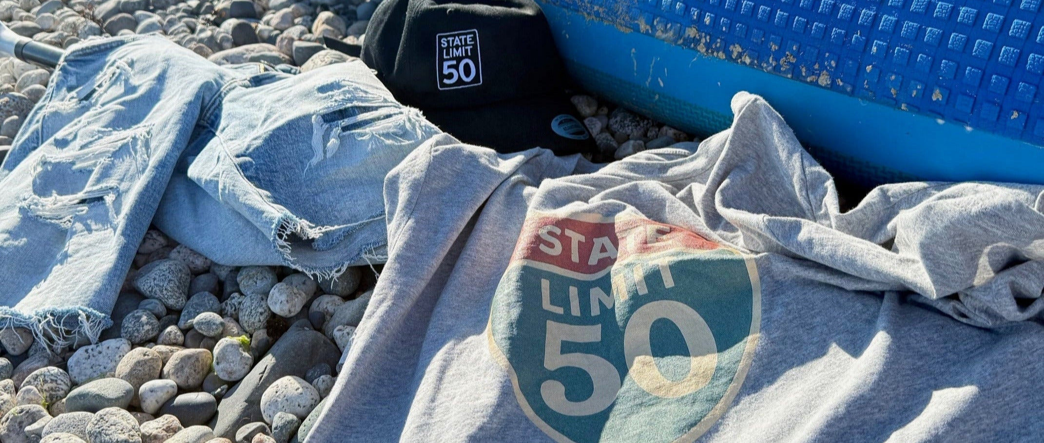 Embroidered hat & graphic tee by State Limit Apparel in the sun on a pebble beach beside distressed jean shorts & a paddleboard.